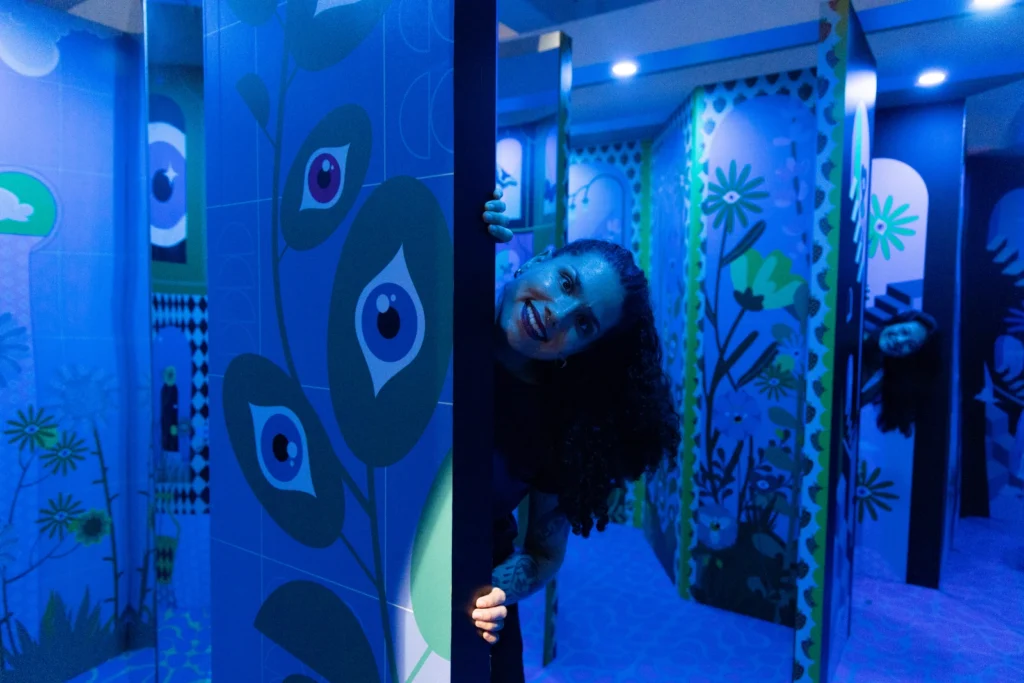 A visitor peeking around a mirrored wall inside a blue-lit floral illusion room at Mind Games Sydney, with repeated reflections creating a playful maze effect.