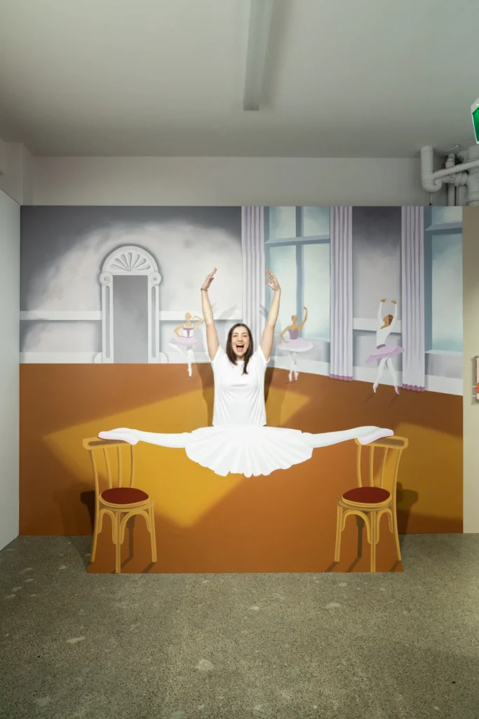 A visitor posing as a ballerina performing a split between two chairs in a playful 3D illusion artwork at Mind Games Sydney.