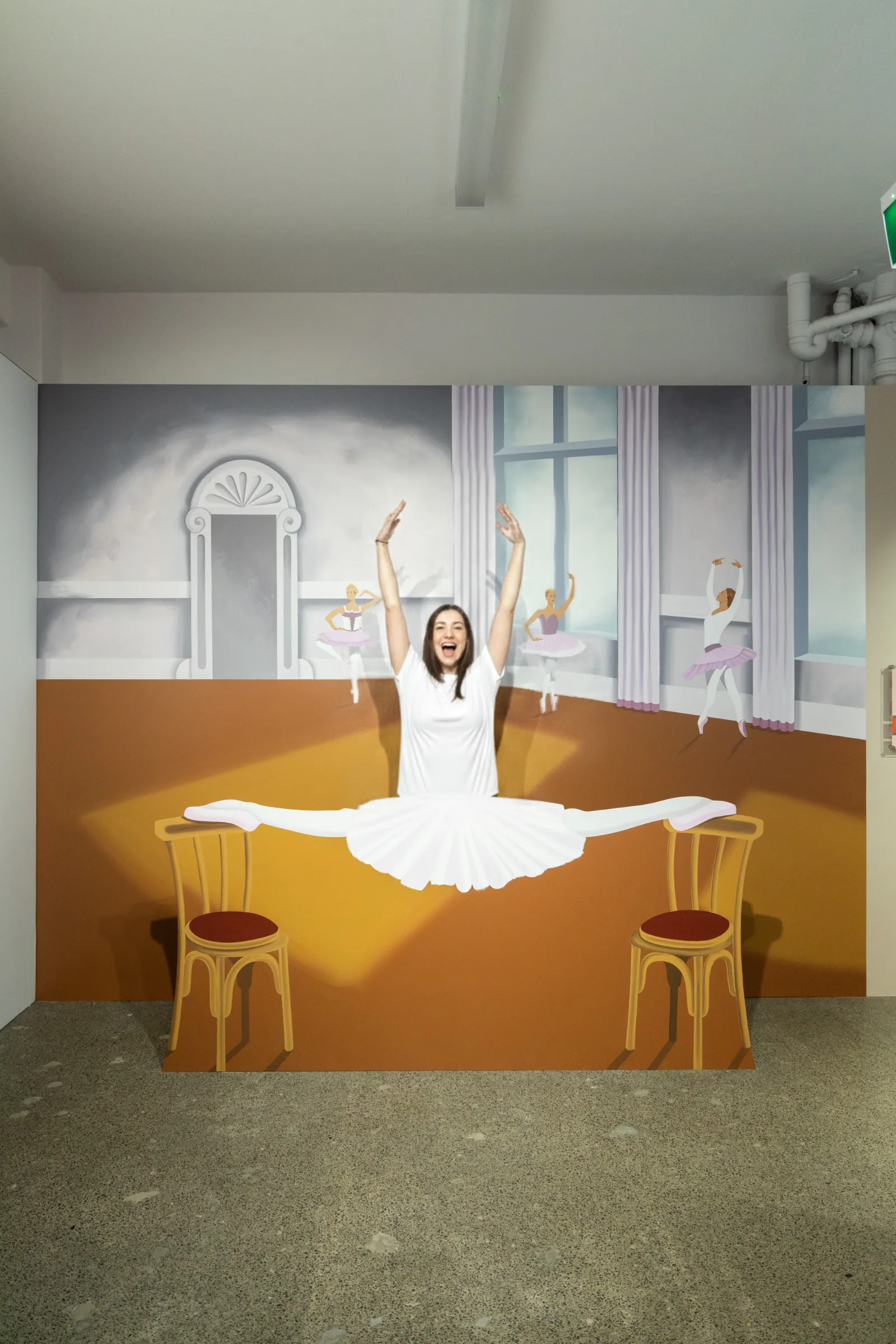 A visitor posing as a ballerina performing a split between two chairs in a playful 3D illusion artwork at Mind Games Sydney.