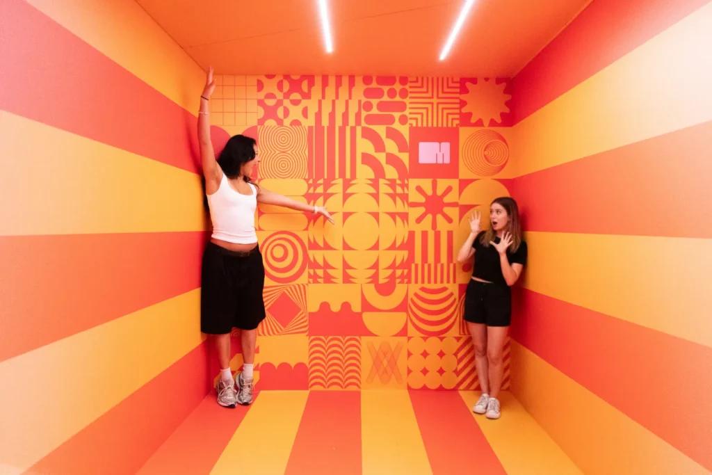 Two visitors posing in a vibrant orange-and-yellow perspective illusion room at Mind Games Sydney, creating the appearance of dramatically different heights.