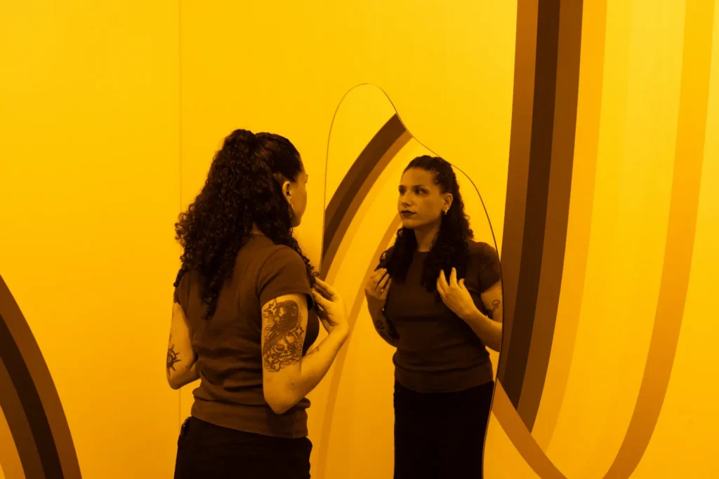 A visitor looking at her reflection in a curved mirror inside a yellow optical illusion room at Mind Games Sydney.