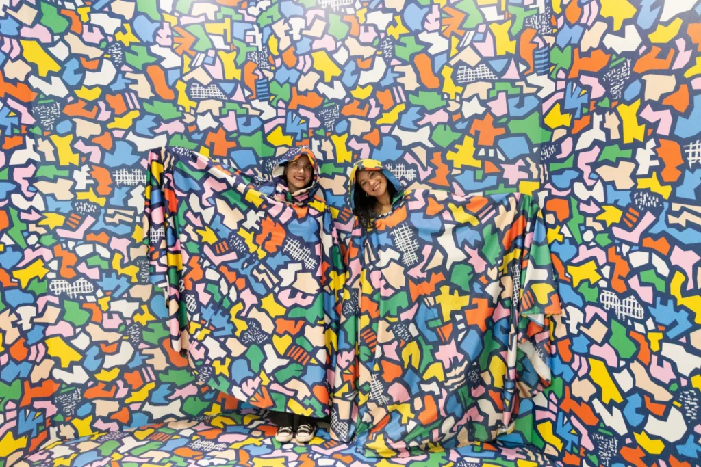 Two visitors blending into a colourful camouflage room at Mind Games Sydney, wearing matching patterned fabric against an abstract multicoloured wall.