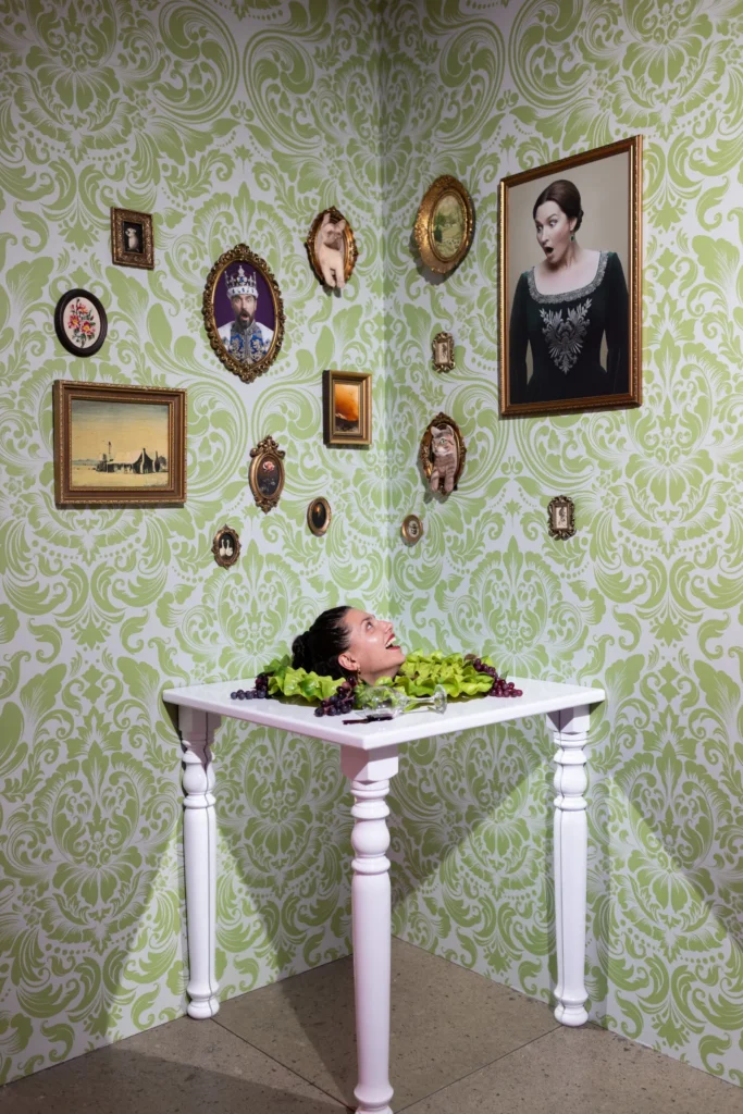 A visitor posing with her head appearing through a dining table in a whimsical green vintage illusion room at Mind Games Sydney.