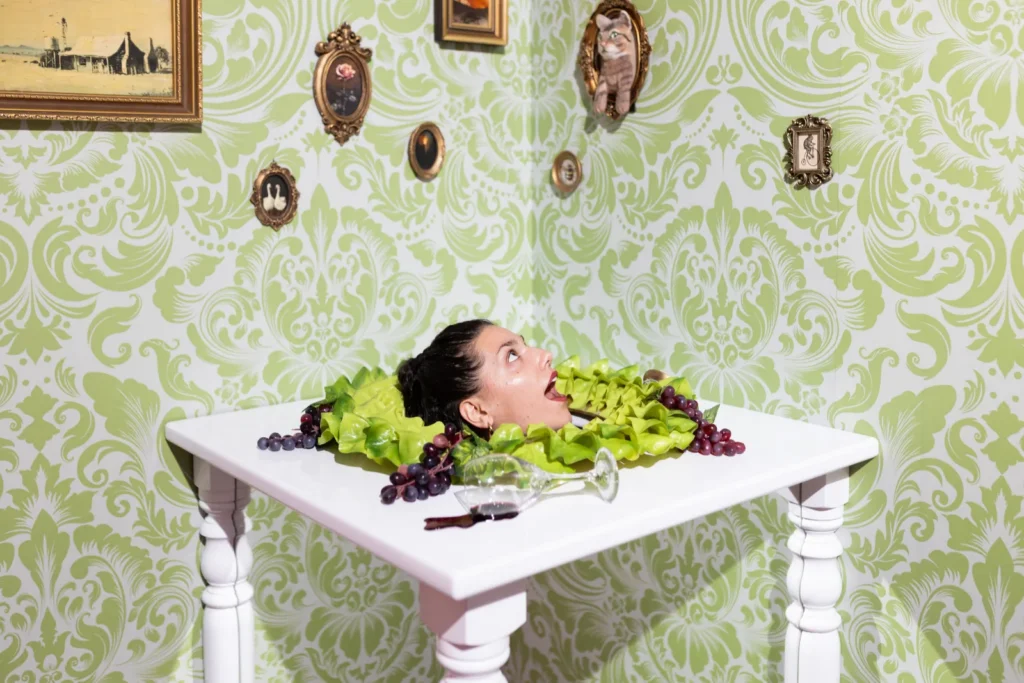 A visitor posing with her head appearing through a dining table in a whimsical green vintage illusion room at Mind Games Sydney.