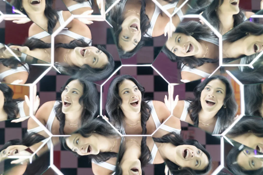 A visitor surrounded by multiple reflections in a geometric kaleidoscope mirror illusion at Mind Games Sydney.