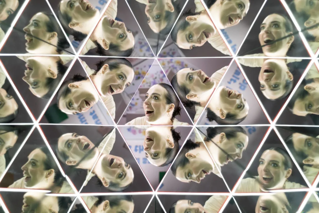 A visitor surrounded by multiple reflections of her face inside a geometric kaleidoscope mirror illusion at Mind Games Sydney.