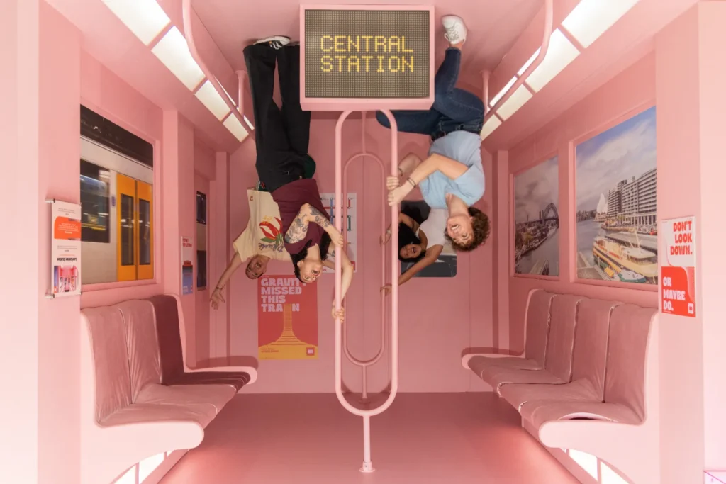 Four visitors posing upside down inside a pink gravity-defying train illusion room at Mind Games Sydney.