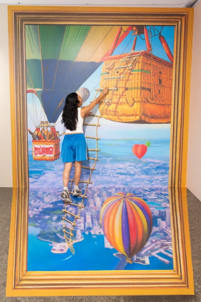 A visitor climbing a rope ladder into a colourful hot air balloon 3D illusion artwork at Mind Games Sydney.
