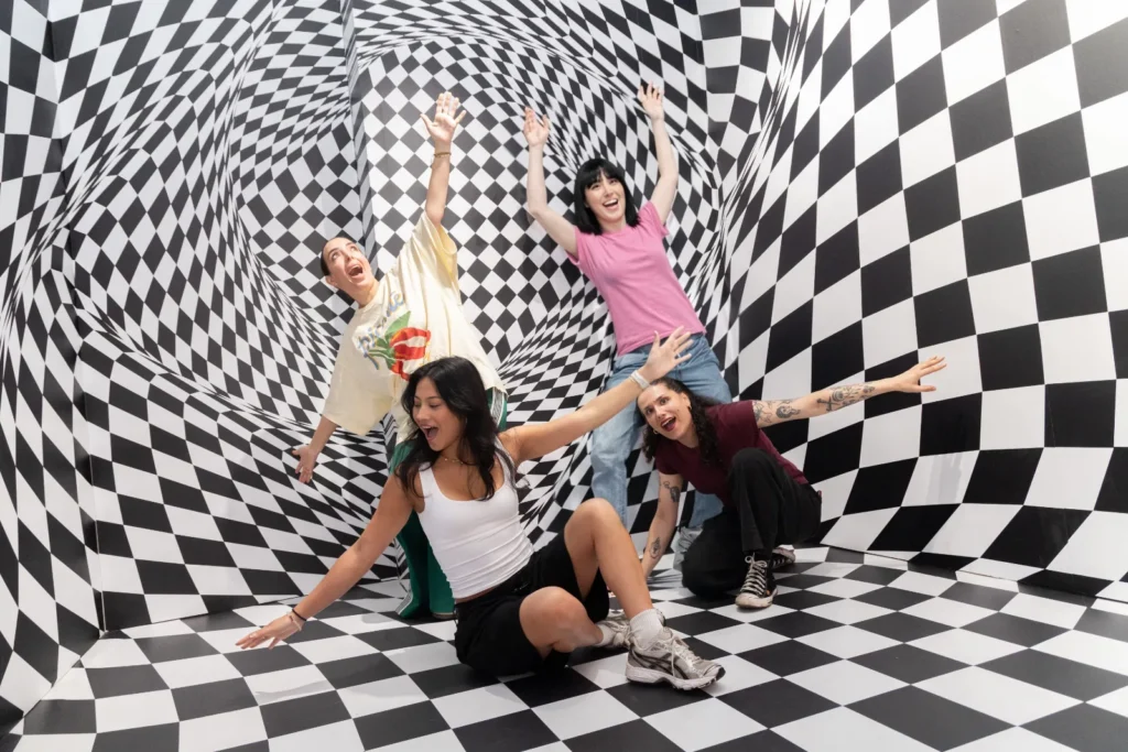Four visitors posing inside a black-and-white checkerboard illusion room at Mind Games Sydney, surrounded by warped geometric patterns that create a distorted perspective effect.