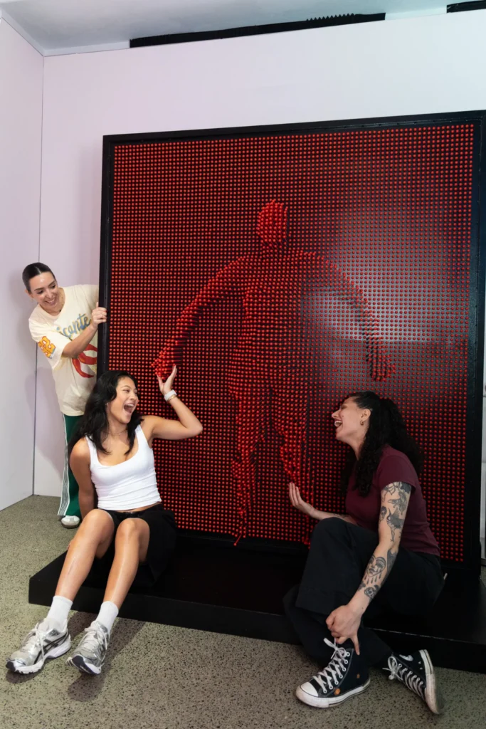 Three visitors laughing and interacting with a large red pin wall installation at Mind Games Sydney, with a human silhouette pressed into the pins.