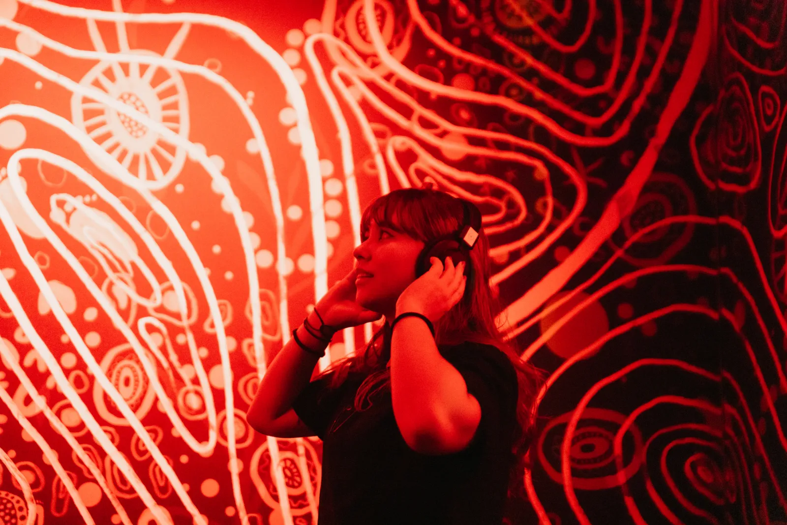 A visitor wearing headphones experiences an immersive red-lit artwork with glowing hand illustrations at Mind Games Sydney.